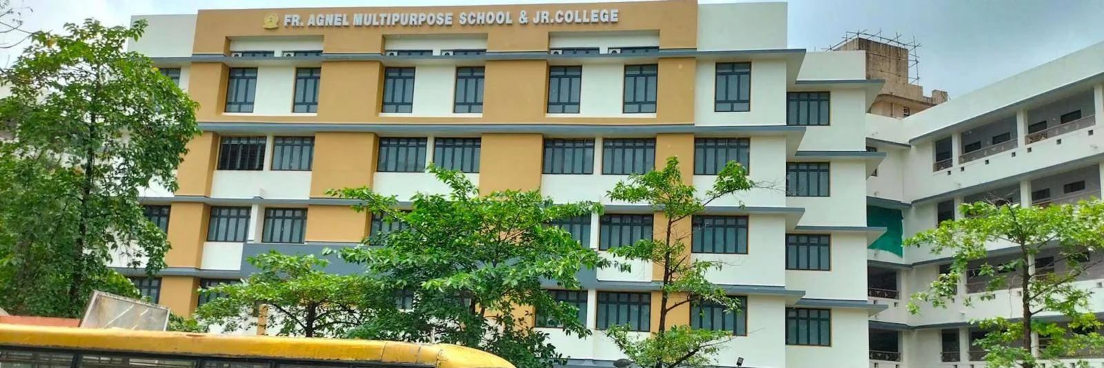 The school and Jr. College building: a multi-storey block in cream and ochre bands with the sign “Fr. Agnel Multipurpose School & Jr. College” across the top, trees in front and a yellow school bus along the bottom edge.