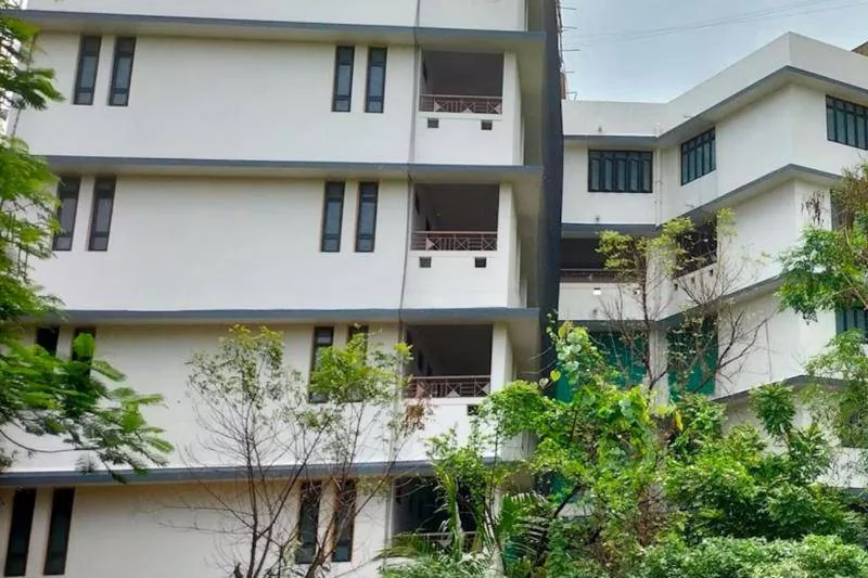 A white wing of the school with narrow windows, open balconies and green trees growing against the walls.