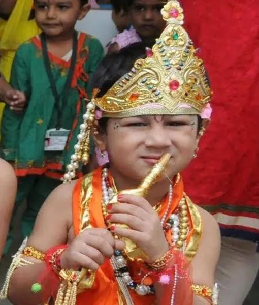 A child in an orange costume and tall jewelled crown holding a golden flute to their lips, with other children behind.