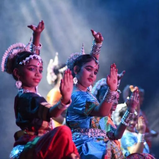 Children in classical dance costumes with raised hands and ornate headdresses, lit by blue stage lights.