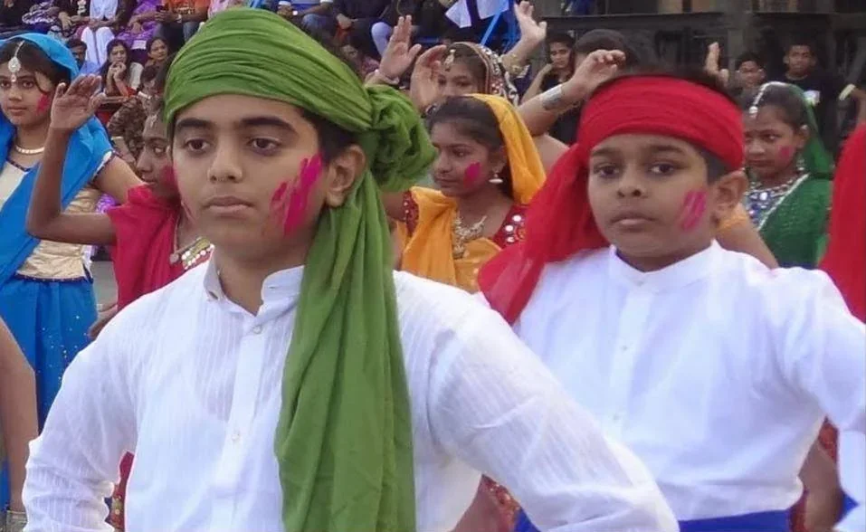 Two children in white shirts, one in a green turban and one in a red headband, with colour on their cheeks, performing a folk dance in front of other costumed children.