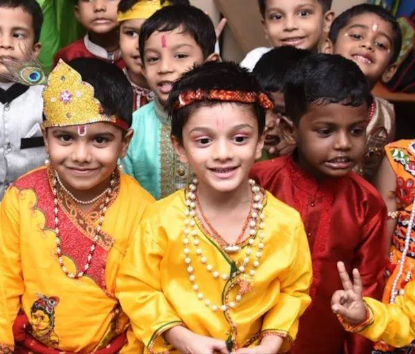 Smiling children in yellow and red kurtas with beaded necklaces and decorated headbands, standing close together.
